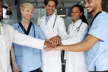 Takım çalışması, işbirliği konsepti. Profesyonel doktorlardan oluşan olumlu, çok ırklı bir ekip. İş elbiselerindeki erkekler ve kadınlar neşeleniyor, ellerini birbirinin üstüne koyuyor ve gülümsüyor, yakın plan fotoğraf çekiyorlar.