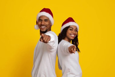 Mutlu çift sırt sırta pozlar veriyor, ikisi de Noel Baba şapkası ve beyaz gömlek giyiyor. Kamerayı gösteriyorlar, canlı sarı bir ortamda gülüşlerini paylaşıyorlar. Tatil ruhu için mükemmel..