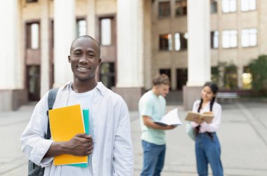 Bir grup üniversite öğrencisi kampüste birlikte çalışıyor. Bir öğrenci elinde not defterleri tutarken gülümserken, iki akran arka planda konuşuyorlar ve materyalleri gözden geçiriyorlar.