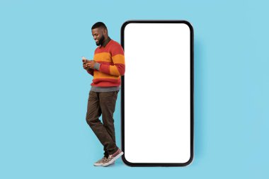 Mobile Offer. Smiling Blank Man With Smartphone Standing Near Big Blank Telephone With White Screen, Happy Young African American Male Enjoying New Application Or Website, Collage, Mockup