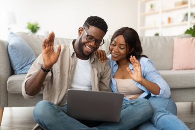 Siyahi bir çift yerde oturup video konferansı sırasında dizüstü bilgisayarlarına gülümseyip el sallıyor..