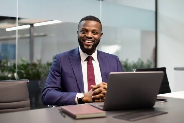 Modern ofisteki çalışma masasında poz veren takım elbiseli neşeli, yakışıklı genç siyahi iş adamı, dizüstü bilgisayarın önünde oturan Afro-Amerikan girişimci, kameraya gülümseyen, uzay kopyası