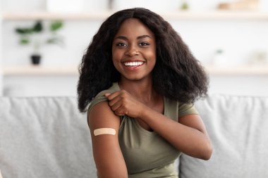 Genç bir kadın kanepede oturuyor, kolundaki bandajı gösterirken gülümsüyor. Evinde sıcak bir ortamda aşı olduktan sonra mutlu görünüyor..