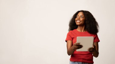 Neşeli siyah bir kadın iki eliyle dijital bir tablet tutuyor, ilgiyle yana bakıyor. Sade beyaz bir geçmişi olan parlak bir stüdyoda neşeli bir atmosfer yaratıyor..
