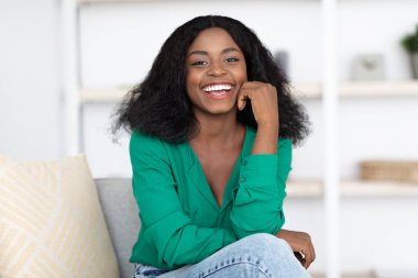 Uzun, siyah saçlı bir kadın, aydınlık bir odada koltukta otururken neşeyle gülümsüyor. Yeşil bir bluz giyer ve eli çenesinde rahatça poz verir..