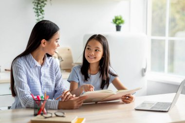 Sıcak bir mutfakta, Koreli bir anne kızına ödevlerinde yardım eder. Gülümseyen kız, sıcak ve davetkar bir atmosferle çevrili neşeli bir anı paylaşırken bir kitap tutuyor..