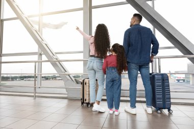 Bir aile havaalanı terminalinde duruyor, büyük pencerelerden dışarı bakıyor. Bagajları var ve yaklaşan yolculukları için heyecanlı bir uçağın gökyüzüne çıkışını izliyorlar..