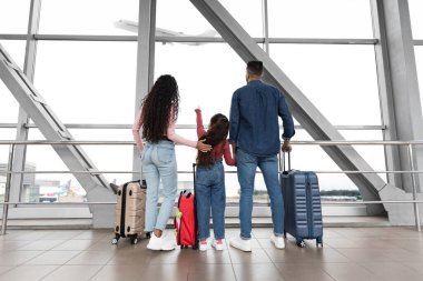 Ailesi bir havaalanının içinde yukarıda uçan bir uçağa bakıyor. Yaklaşan yolculuk için heyecanlı ve hazır görünüyorlar, yanlarında bavullarıyla..