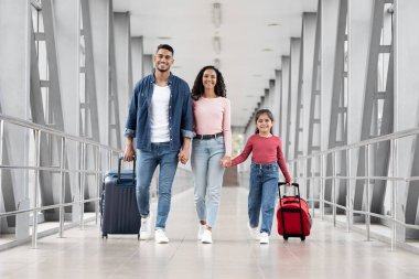 Üç kişilik bir aile, parlak bir havaalanı terminalinden el ele geçiyor. Renkli bavullar taşıyorlar ve uçuşlarına hazırlanırken neşeli anları paylaşıyorlar..