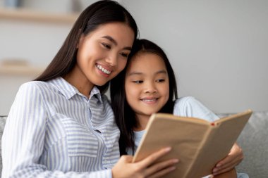 Neşeli bir Asyalı anne ve tatlı kızı büyüleyici bir kitaba dalmış bir kanepede birlikte oturuyorlar. Evde geçirdikleri kaliteli zaman boyunca güzel bir hikayenin tadını çıkarırlarken aynı gülümsemeyi paylaşırlar..