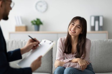 Neşeli bir Arap kadın kanepede oturup rahat bir klinikte psikoloğuyla sohbet ediyor. Seanslarında duygularını ve deneyimlerini tartışırlarken gülümsüyor..