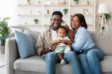 Neşeli siyah baba ve anne, sevimli oğullarıyla parlak bir oturma odasında poz veriyorlar. Aile bir kanepede oturur, gülümser ve evde sevgi dolu bir anı paylaşır..
