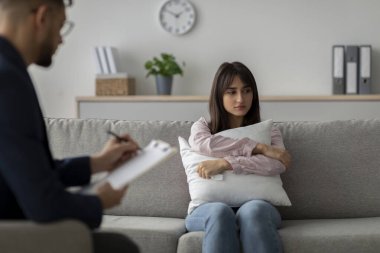 Orta Doğulu bir kadın terapist ofisindeki bir kanepede oturur ve yastığa sarılırken mutsuz görünür. Psikoterapist sakin bir ortamda dikkatlice dinlerken notlar alır..