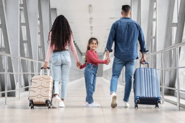 Üç kişilik mutlu bir aile havaalanı terminalinde el ele yürüyor. Aileler, neşeli kızlarına gülümserken bavullarını taşıyorlar. Yolculukları için heyecanlılar..