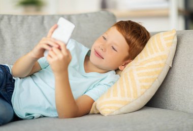 Neşeli bir kızıl saçlı çocuk evdeki rahat bir kanepede uzanıyor, akıllı telefonunu arkadaşlarıyla sohbet etmek, oyunlar oynamak ve yeni uygulamalar keşfetmek için kullanıyor. Yüzündeki ifade neşe ve bağlılık gösteriyor..