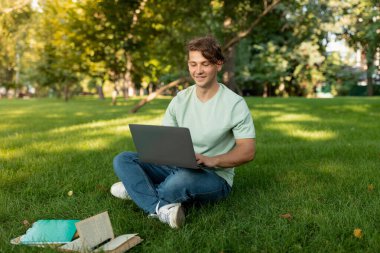 Genç bir adam çimenlerde bacak bacak üstüne atarak otururken gülümsüyor, güneşli bir parkta dizüstü bilgisayarına odaklanıyor. Etrafında ağaçlar ve üretkenlik için mükemmel sakin bir atmosfer var..
