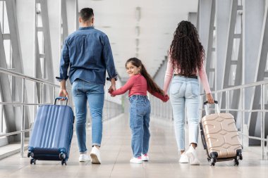Bir aile, parlak bir havaalanı terminalinde el ele tutuşarak yürür. Anne bej bir çanta taşırken baba mavi bir bavul taşıyor. Aralarında genç bir kız gülümsüyor..