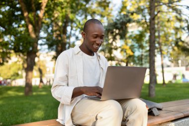 Genç bir adam güneşli bir parkta ahşap bir bankta dizüstü bilgisayar kullanırken gülümsüyor. Ağaçlar onu çevrelemiş, iş ve eğlence için huzurlu bir ortam yaratmış..