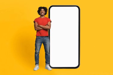 Kıvırcık saçlı genç bir adam kolları çaprazlanmış şekilde büyük, boş bir akıllı telefon çerçevesinin yanında poz veriyor. Parlak sarı arka plan, akıllı telefonların varlığını vurgulayarak sahneye canlı bir dokunuş katıyor..