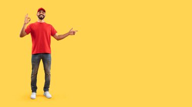 Kırmızı t-shirt ve kot giyen neşeli bir adam, parlak sarı bir arka plana karşı duruyor. Gülümsüyor ve olumlu bir işaret veriyor. Olumlu bir şey söylüyor..