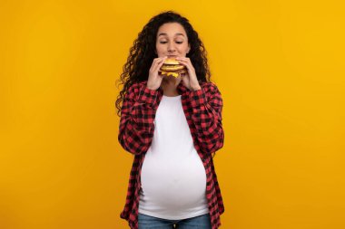 Neşeli hamile bir kadın abur cubura olan aşkını gösteren büyük bir hamburgerin tadını çıkarıyor. Canlı sarı-turuncu bir arka plana karşı poz veriyor, hoşgörü dolu anını vurguluyor..