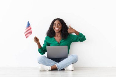 Genç bir kadın bir elinde küçük bir Amerikan bayrağını tutarken, diğer elinde başparmağıyla yerde sırıtarak oturuyor. Parlak bir alanda dizüstü bilgisayar kullanıyor..