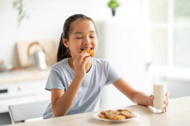 Neşeli bir kız mutfak masasında oturur, elinde bir bardak süt tutarken mutlu bir şekilde lezzetli bir kurabiye yer. Gülümsemesi, evdeki sağlıklı atıştırmalıkların keyfini gösteriyor..