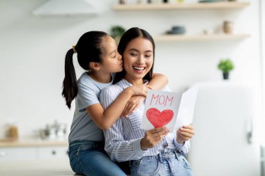 Küçük bir kız, büyük kırmızı bir kalple süslenmiş bir hediye kartıyla annesini mutlu bir şekilde karşılıyor. Annesini yanağından öpüyor, sevgisini ve minnettarlığını ifade ediyor..