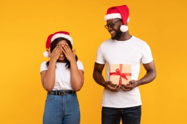 Noel Baba şapkalı Afrikalı bir adam bir kadını şaşırtırken sarılı bir hediye tutuyor. Gözlerini kapatıyor, heyecanlı, neşeli sarı stüdyo ortamında tatil sezonunun neşesini yakalıyor..