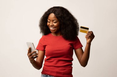 Neşeli bir Afrikalı Amerikalı kadın beyaz bir arka plana karşı duruyor, akıllı telefonunu online alışveriş yapmak için kullanırken gülümsüyor. Elinde dijital ödemelerin kolaylığını gösteren bir kredi kartı var..