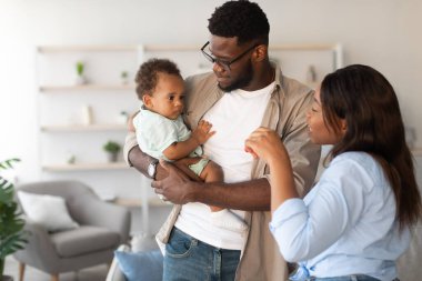 Genç Afrikalı Amerikalı ebeveynler keyifle sevimli bebekleriyle sıcak bir oturma odasında etkileşim halindeler..
