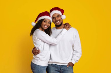 Mutlu bir çift Noel Baba şapkaları ve rahat kazaklar giyerek birbirlerini kucaklıyor. Neşeli sarı arka plan, bayram ruhunu ve o anın neşesini arttırır..