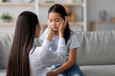 Genç bir Asyalı anne evdeki bir kanepede oturuyor ve anlaşmazlıktan sonra onu sakinleştirmek için ellerini kızlarının yüzüne koyuyor. Atmosfer anlayış ve desteği yansıtır..