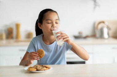 Genç kız mutfak masasında oturuyor, süt yudumluyor ve kurabiyelerin tadını çıkarıyor. O, temiz ve parlak bir mutfak alanı ile çevrili atıştırmalığının keyfini çıkarırken mutlu görünüyor..