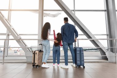 Mutlu bir aile havaalanının içinde, gökyüzündeki bir uçağa büyük pencerelerden bakıyor. Çocuk heyecanlı bir şekilde yolculuklarına hevesli olduğunu gösteriyor..