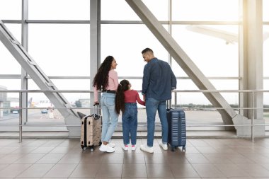 Havaalanı terminalinde bir aile bir arada duruyor. Dışarıdaki uçaklara büyük pencerelerden bakıyorlar..