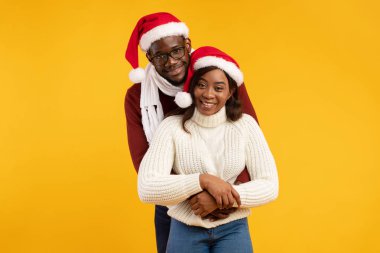 Noel Baba şapkası takan neşeli siyah bir çift, canlı sarı bir zemin karşısında yan yana duruyorlar. Gülümsüyorlar ve Noel ruhunu kutluyorlar. Sevgi ve mutluluk gösteriyorlar..