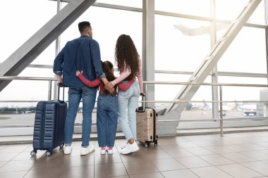 Bir aile havaalanı terminalinde durup dışarıdaki uçaklara büyük pencerelerden bakıyor. Yolculuklarına hazırlanırken heyecanları ve beklentileri çok açık..
