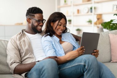 Neşeli bir Afrikalı Amerikalı çift, rahat ve bağlı bir şekilde koltuklarında oturuyor. Tabletlerine bakarken kahkahaları paylaşıyorlar, rahat oturma odalarında videoların ve anıların tadını çıkarıyorlar..