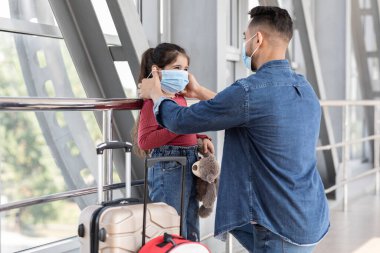 Bir baba havaalanında kızının maskesine yardım etmek için diz çöker. Bir bavulun yanında durup yolculuklarına hazır olduklarını ve güvenlik uygulamalarına özen gösterirler..