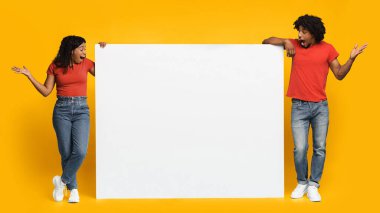 İki kişi büyük boş bir tahtanın her iki tarafında sarı bir zemin üzerinde duruyor. Sıradan kırmızı gömlekler ve kotlar giyiyorlar, heyecan ve sürpriz sergiliyorlar..