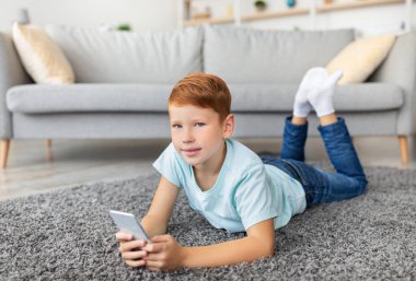 Sıcak bir oturma odasında yerde yatan kızıl saçlı neşeli bir genç çocuk. O, cep telefonuyla meşgul, arkadaşlarıyla sohbet ediyor ve oyunlardan zevk alıyor. Yumuşak güneş ışığı boşluğu doldurur.