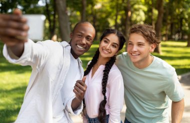 Üç arkadaş gülümser ve güneşli bir parkta selfie için poz verir. Kahkaha ve dostlukla dolu neşeli bir anı yakalarken parlak yeşil ağaçlar etraflarını sarar..