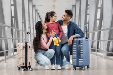 Üç kişilik neşeli bir aile havaalanında mutlu bir anı paylaşıyor. İki bavulu var ve bir çocuk oyuncak ayı tutuyor, gülümsüyor ve birbirlerine sarılıyorlar..