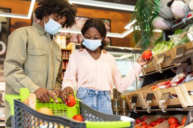 Bir çift, her biri maske takan domatesleri marketten seçiyor. Alışveriş arabalarını taze meyve ve sebzelerle doldururken odaklanmış görünüyorlar..