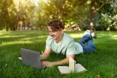 Güneşli bir parkta, genç bir adam çimlere uzanır, dizüstü bilgisayarına odaklanır. Sıcak bir günde dışarıda verimli bir çalışma seansının keyfini çıkarırken kitaplar onun yanında..