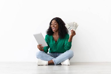 Neşeli bir kadın bağdaş kurup yerde oturuyor, bir elinde para, diğer elinde tablet var. Parlak bir odada, temiz, minimalist bir tasarımı var..