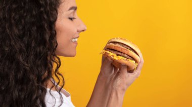 Gülümseyen bayan sarı turuncu bir duvarla stüdyoda duruyor ve büyük lezzetli burgerini göstermeye can atıyor. Lezzetli sandviçe olan heyecanı kameraya poz verirken açıkça görülüyor..