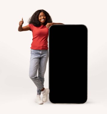 Neşeli siyah bayan büyük boş bir akıllı telefonun yanında duruyor. Parlak beyaz stüdyo arka planında bir mobil uygulamayı veya web sitesini tanıtıyor..
