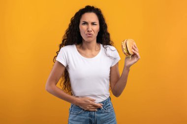 Üzgün bir Latin kadın elinde hamburgerle sarı turuncu bir arka plana karşı duruyor. Sağlıksız bir yemek seçiminden sonra karnına dokunuyor ve rahatsızlık veriyor..